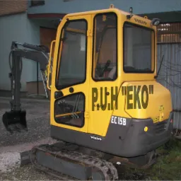 Żółta minikoparka Volvo EC 15B z czarnym napisem 'P.U.H 'EKO'' na karoserii, stojąca na trawie przed budynkiem.